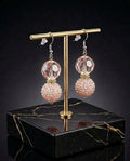 A pair of dangling earrings displayed on a gold-tone stand over a black marble base. Each earring has a faceted pale pink crystal ball at the top and a textured, rhinestone-covered pink sphere below.