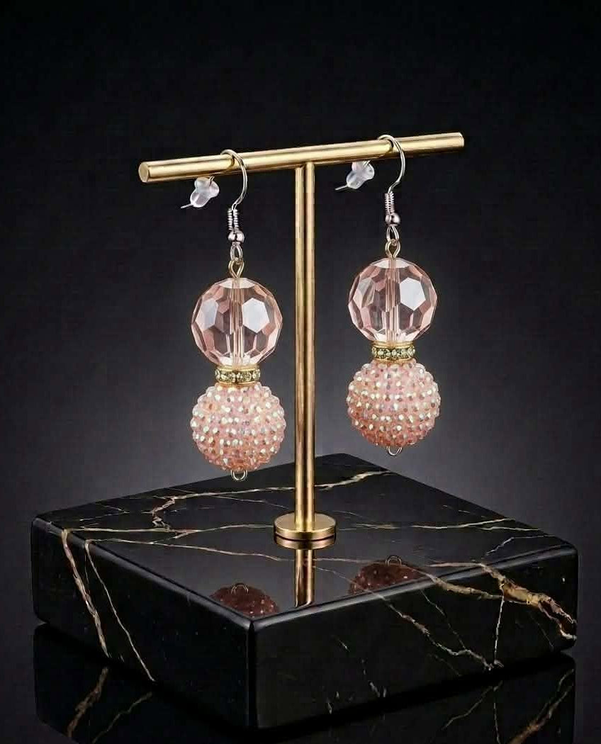 A pair of dangling earrings displayed on a gold-tone stand over a black marble base. Each earring has a faceted pale pink crystal ball at the top and a textured, rhinestone-covered pink sphere below.